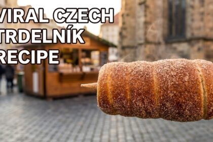 A close-up shot of a golden-brown Trdelník (chimney cake) coated in cinnamon sugar and walnuts, standing upright on a rustic wooden table in a 2026 kitchen.