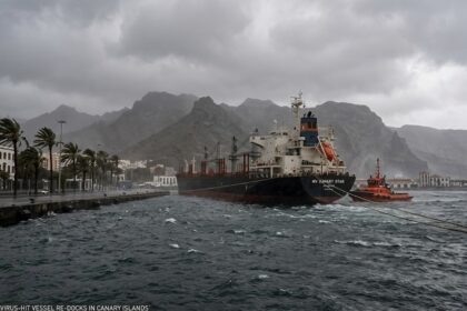 A digital graphic showing the MV Hondius moored in the Port of Granadilla under heavy winds, with medical staff in biohazard suits standing near an ambulance.